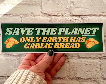 May include: A green sticker with the text "SAVE THE PLANET" and "ONLY EARTH HAS GARLIC BREAD" in white and orange. The sticker features illustrations of garlic bread slices. The sticker is held by a hand.