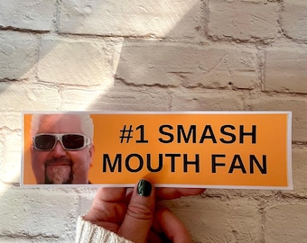 May include: An orange rectangular sticker with the text "#1 SMASH MOUTH FAN" in black. The sticker features a picture of a man wearing sunglasses. The sticker is held by a person with a cream-colored sweater.