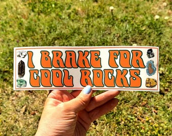 May include: A rectangular sticker with the text "I Brake For Cool Rocks" in orange bubble letters. The sticker has illustrations of various rocks and crystals. The sticker is held in front of a grassy background.