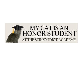 May include: A rectangular sticker with the text "MY CAT IS AN HONOR STUDENT AT THE STINKY IDIOT ACADEMY." A gray cat wearing a black graduation cap with a yellow tassel is on the left side of the sticker.