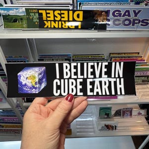 May include: A black bumper sticker with the text "I BELIEVE IN CUBE EARTH" in white. The sticker features a graphic of Earth as a cube. Other bumper stickers are visible in the background.