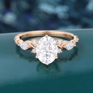May include: A rose gold ring featuring a large oval-cut center stone, flanked by two smaller pear-cut side stones and several tiny round accent stones. The band has a twisted, textured design that leads up to the central stone setting.