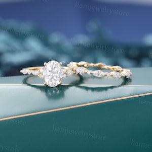 May include: A set of two gold rings. The engagement ring features a large oval-cut diamond center stone, surrounded by smaller diamonds. The wedding band has a delicate design with marquise-shaped stones. The rings are set against a teal and blue background.