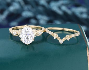 May include: A gold engagement ring set featuring a large oval-cut center stone flanked by smaller marquise-cut stones, paired with a matching V-shaped gold wedding band adorned with a series of tiny round-cut accent stones.