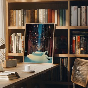 May include: A framed art print depicting a snow-covered street scene at night, with warm lights strung overhead. The print is displayed on a wooden desk with books, a fan, and a small speaker. Bookshelves filled with books are in the background.