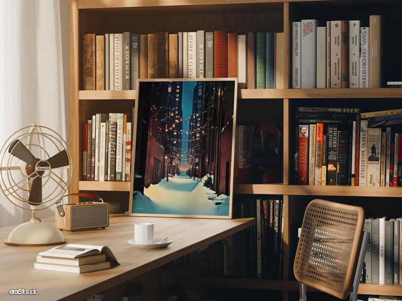 May include: A framed art print depicting a snow-covered street scene at night, with warm lights strung overhead. The print is displayed on a wooden desk with books, a fan, and a small speaker. Bookshelves filled with books are in the background.