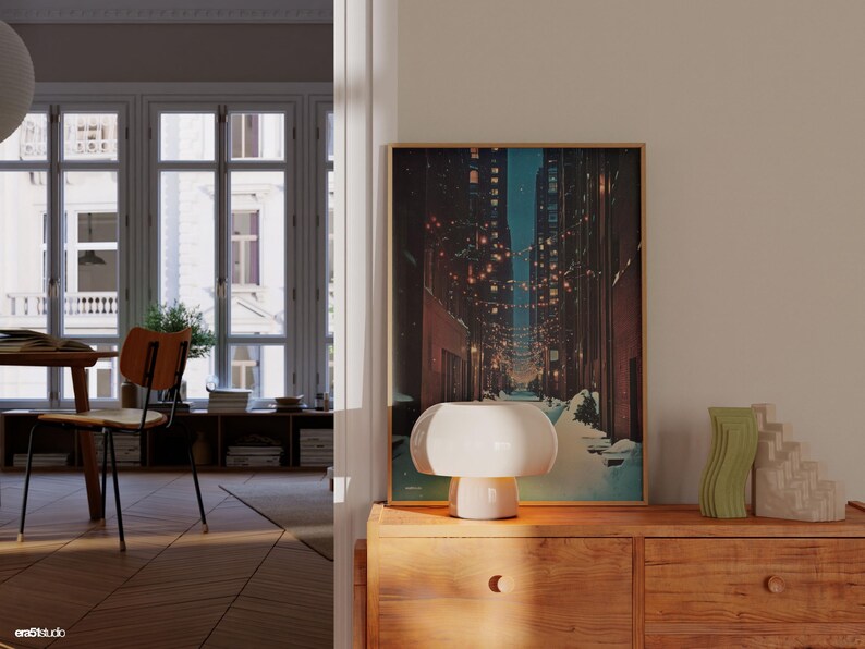May include: A warm-toned interior scene featuring a wooden dresser with a framed artwork depicting a city street at night. A white mushroom-shaped lamp sits on the dresser, alongside decorative vases. The room has a classic design.