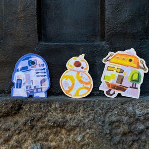 May include: Three die-cut stickers featuring stylised illustrations of science fiction robots. The left sticker is blue and white, the centre sticker is orange and white with a spherical body, and the right sticker is orange, yellow, and green.