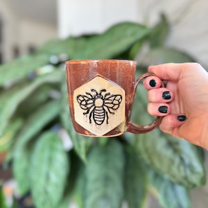 May include: A handmade ceramic mug with a brown exterior and a beige hexagon design featuring a black bee illustration. The mug has a brown handle and is held in front of a blurred green plant background.