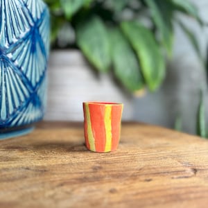 May include: A small, handmade ceramic cup with a vibrant design. The cup features vertical stripes of bright orange and yellow. The cup is set on a wooden surface, with a blue ceramic pot and green plants in the background.