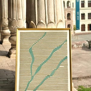 May include: A rectangular framed artwork featuring a series of horizontal gold lines set against a white background. Overlaid on the gold lines are three distinct, flowing teal-colored shapes that resemble organic, branching forms or river-like paths.