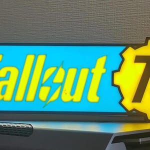 Könnte beinhalten: Ein beleuchtetes Schild mit dem Fallout 76-Logo. Das Schild hat ein rechteckiges blaues Feld mit den gelben Worten "Fallout 76" und ein gelbes Zahnrad-Emblem mit der Zahl "76".