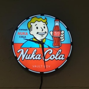 Könnte beinhalten: Runde, beleuchtete Wanduhr mit dem Nuka Cola-Logo und der Figur Vault Boy. Das Zifferblatt ist in blaue und orangefarbene Abschnitte unterteilt, mit den Wörtern "Vintage Nuka Cola" und "Nuka Cola" deutlich sichtbar. Die Uhrzeiger sind schwarz.