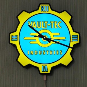 Könnte beinhalten: Eine Wanduhr mit einem gelben, zahnradförmigen Rahmen und einem blauen Zifferblatt. Das Zifferblatt zeigt den Text "VAULT-TEC INDUSTRIES" in Gelb mit einem stilisierten Logo. Römische Ziffern markieren die Stunden. Die Uhr wird von einem weißen Kabel mit Strom versorgt.