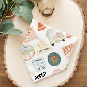 May include: A white dog bandana with colorful camping-themed patches. Patches include a camper van, a campfire, and the words "Explore," "Adventure," and "Wilderness." The name "Aspen" is printed on a small white tag.
