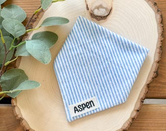 May include: Blue and white striped dog bandana with a white label that says "Aspen" on it. The bandana is on a wooden slice.