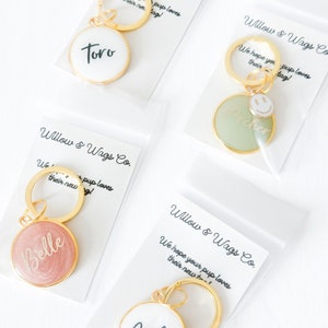 May include: Four gold keychains with round, personalized tags. The tags are white, green, pink, and white with black lettering. The tags say "Toro", "Archer", "Belle", and "Archie".