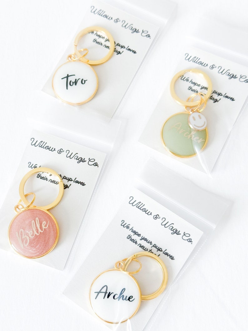 May include: Four gold keychains with round, personalized tags. The tags are white, green, pink, and white with black lettering. The tags say "Toro", "Archer", "Belle", and "Archie".