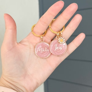 May include: Two pink round keychains with gold rings. One keychain has the name "Mocha" written on it and the other has the name "Iris" written on it. Each keychain has a small white daisy charm.