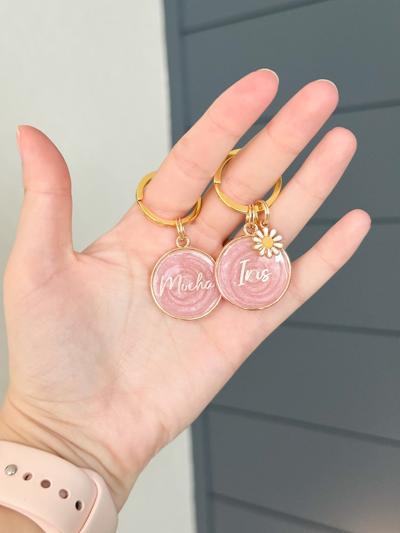 May include: Two pink round keychains with gold rings. One keychain has the name "Mocha" written on it and the other has the name "Iris" written on it. Each keychain has a small white daisy charm.
