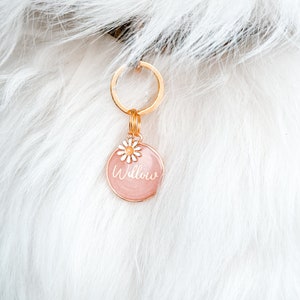 May include: A pink round dog tag with a gold chain and a small white flower charm. The tag says "Willow" in white script.