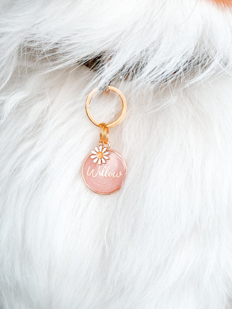 May include: A pink round dog tag with a gold chain and a small white flower charm. The tag says "Willow" in white script.