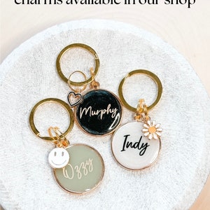 May include: Three gold keychains with round charms. The charms are black, white, and green. The black charm has the name "Murphy" written in white. The white charm has the name "Indy" written in black. The green charm has the name "Izzy" written in gold and a white smiley face charm.