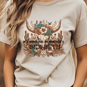 May include: A beige t-shirt with a floral design featuring a skull with horns. The text "In a world full of princesses, be a Beth" is printed on the shirt.