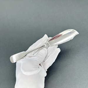May include: A silver butter knife with a curved, scalloped blade and a long, slender handle. The handle has a rectangular end with an engraved initial. The knife is held by a gloved hand against a gray background.