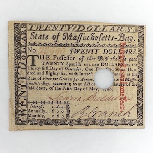 May include: A rectangular historical paper currency note from the State of Massachusetts-Bay, valued at Twenty Dollars. The off-white paper features black printed text, decorative borders, a circular hole, and red ink text reading "To counterfeit is death."