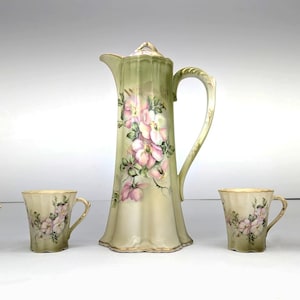 May include: A porcelain tea set consisting of a tall, fluted pitcher and four matching cups. Each piece features a pale green and cream gradient background with hand-painted pink floral motifs and green leaves, finished with gold trim.