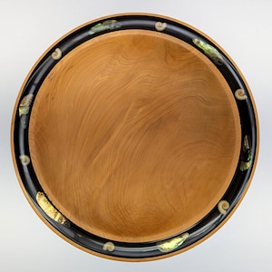 May include: A circular wooden bowl with a smooth, light brown interior featuring visible wood grain. The wide, dark black rim is inlaid with iridescent abalone shell fragments and small, spiral-shaped seashells arranged in a repeating pattern.