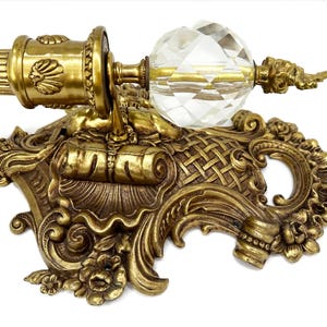 May include: Ornate antique brass curtain rod hardware with a faceted crystal ball finial. The rod features detailed relief carvings and a decorative bracket with floral and scrollwork elements. The overall design is vintage and elegant.