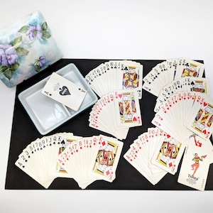 May include: A set of playing cards arranged in four suits on a black surface, with a light blue ceramic tray containing an ace of spades card and a matching floral patterned ceramic box lid featuring purple flowers and green leaves.