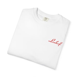 May include: A folded white t-shirt with a red "Linhof" script logo on the front. The shirt has a crew neck and a tag visible at the neckline. The shirt is made of a soft, comfortable material.