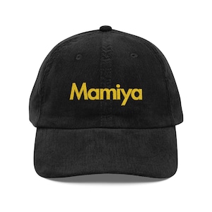 May include: Black corduroy baseball cap with the word "Mamiya" embroidered in gold lettering across the front. The cap has a curved brim and a rounded crown. A classic, casual accessory.