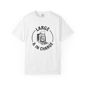 May include: White t-shirt with a black graphic of a vintage camera inside a circle, with the words "LARGE & IN CHARGE" around it. The shirt has a crew neck and short sleeves.