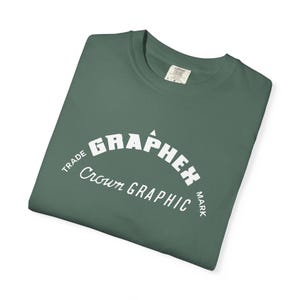 May include: Folded forest green t-shirt with a white graphic that reads "GRAPHEX Crown GRAPHIC" in an arched design. The shirt has a crew neck and a small tag on the collar. The shirt is made of a soft, comfortable material.