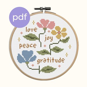 May include: A cross-stitch pattern in a wooden hoop features three flowers in pink, yellow, and blue, with the words "love," "joy," "peace," and "gratitude." The pattern includes green leaves and small star accents, with a "pdf" icon.