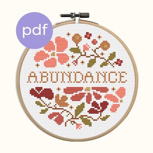 Puede incluir: Un bastidor de bordado de punto de cruz con la palabra "ABUNDANCE" bordada en hilo marrón. El diseño incluye elementos florales en tonos rojos, rosas y verdes. Un círculo morado con la palabra "pdf" está en la esquina superior izquierda.