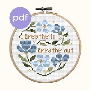 Peut inclure: Une broderie au point de croix dans un cercle en bois présente des fleurs bleues et des feuilles vertes. Les mots "Breathe in" et "Breathe out" sont brodés en fil marron. Un cercle violet avec "pdf" est en haut à gauche.
