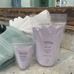 May include: Two bags of lavender milk bath, one large and one small, both with the text "HERITAGE Artisan Soap" and "MILK BATH Lavender Mist" on the front. The larger bag is filled with a purple bath salt and the smaller bag is empty.