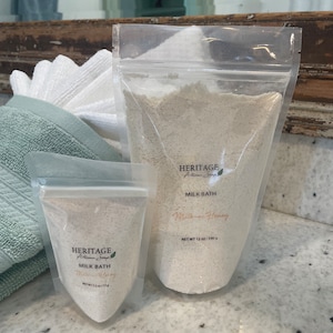 Op de afbeelding: Twee doorzichtige plastic zakjes met Heritage Artisan Soap Milk Bath, Milk & Honey. De grotere zak bevat 340 g product. De kleinere zak bevat 71 g. De zakjes zijn gevuld met een lichtgekleurd poeder.