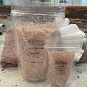 Pode incluir: Duas embalagens de sais de banho, uma grande e uma pequena, ambas com a etiqueta "HERITAGE Artisan Soap Company" e "Cashmere Cream". A embalagem maior é de 20 oz (567 g) e a embalagem menor é de 3,5 oz (100 g).