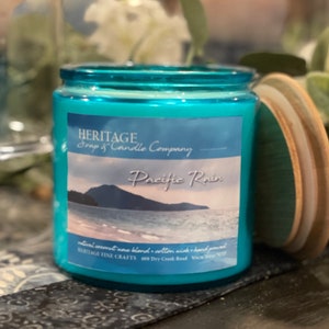 May include: A turquoise glass jar candle with a wooden lid. The candle has a label that reads "Pacific Rain" with a coastal scene. The jar is filled with a light blue wax. The candle is from Heritage Soap & Candle Company.