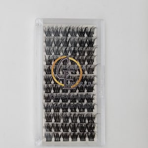 Pode incluir: Uma caixa de plástico transparente contendo vários grupos de cílios postiços pretos. A caixa tem um logotipo dourado e preto que diz "CF COLLECTION". Os cílios são dispostos em fileiras arrumadas.