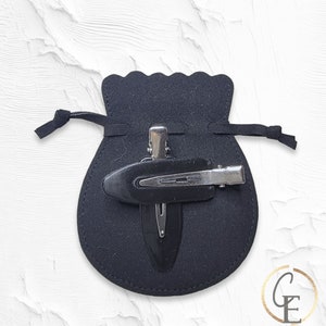 May include: A black velvet pouch with a drawstring closure, containing two black hair clips. One clip is a simple bar clip, and the other is a decorative clip with a teardrop shape.
