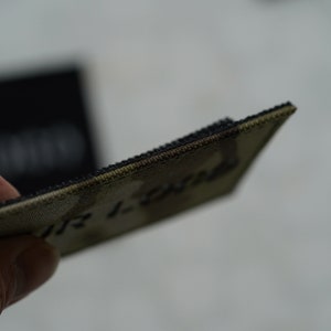 K&ouml;nnte beinhalten: Ein rechteckiger Aufn&auml;her mit einem Tarnmuster in Olivgr&uuml;n- und Braunt&ouml;nen. Der Aufn&auml;her hat einen dicken, dunkel gef&auml;rbten gestickten Rand und eine Klettverschluss-R&uuml;ckseite. Der Text "YOUR LOGO" ist teilweise auf der Oberfl&auml;che zu erkennen.