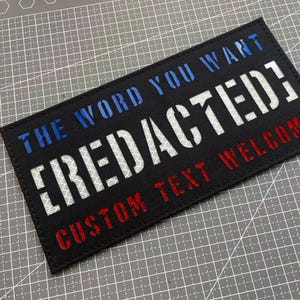 May include: A rectangular black patch with stitched edges featuring three lines of text. The top line reads THE WORD YOU WANT in blue, the middle line reads REDACTED in large white letters, and the bottom line reads CUSTOM TEXT WELCOME in red.
