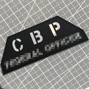 May include: A trapezoidal black fabric patch featuring the reflective silver letters C B P in a bold, sans-serif font. The patch has a stitched border and includes additional blurred text below the main lettering, presented on a grid surface.